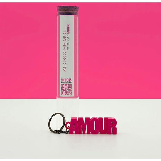 Porte-clés rose AMOUR en lettres majuscules, présenté dans un tube à essai avec bouchon en liège, sur fond rose – Mauvaises Graines
